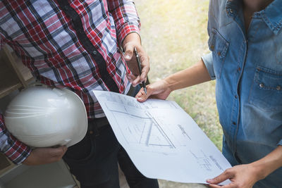 Midsection of female architect holding blueprint while colleague carrying hard hat on field