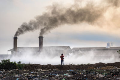 Man standing by smoke emitting from factory against sky