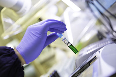 Male doctor's hand wearing a medial glove holding a green vial from a rack in a lab background.