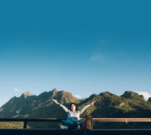 Smiling woman with arms raised sitting on railing with mountains in background