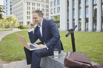 Mature businessman sitting on a wall in the city using laptop