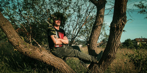 Portrait of smiling man with arms crossed sitting on tree in forest