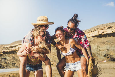 Girlfriends piggybacking boyfriends at beach against sky