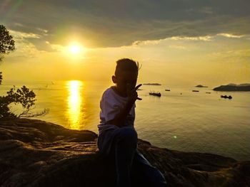 Young man sitting on shore at beach against sky during sunset