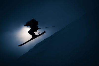 Silhouette of man going off jump with skis and backpack