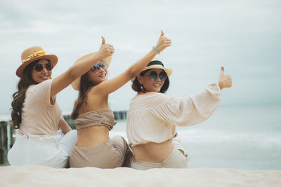 Portrait of women gesturing while sitting at beach against sky