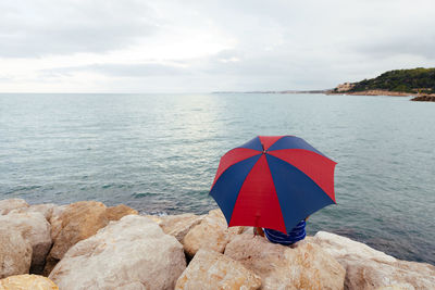 From behind anonymous man sitting on the rocks with an umbrella by the sea while it rains