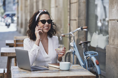 Smiling woman talking on phone while having drink at sidewalk cafe