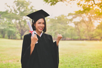 Portrait of young woman in graduation gown holding certificate while standing at park