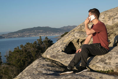 Man sitting on rock by sea against clear sky