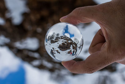 Cropped hand holding crystal ball on landscape during winter with reflection