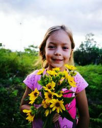 Portrait of happy girl holding yellow flowers while standing on field against sky