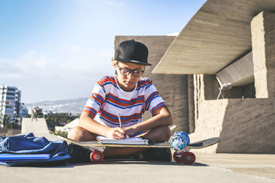 Boy writing on book while sitting outside building