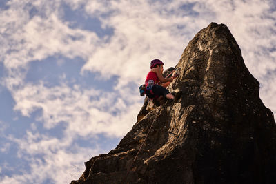 Low angle full length of active senior woman climbing on rocky cliff against sky