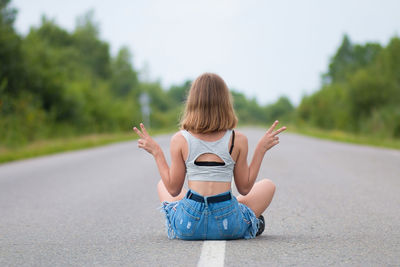 A young girl sits on the road and shows her fingers a sign of victory. summer day