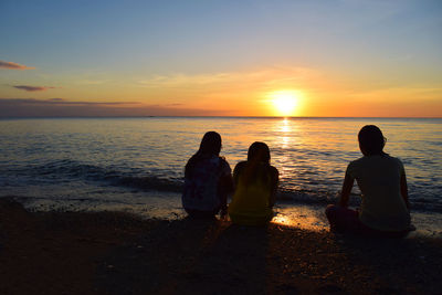 Silhouette friends sitting on beach against sky during sunset