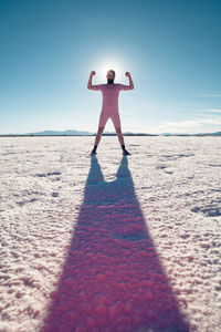 Full length of man flexing muscles while standing on sand at beach against sky during sunny day