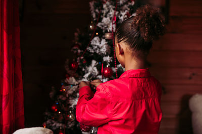 Back view of cheerful little black girl in red shirt looking at camera and smiling while standing near decorated christmas tree at home