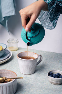 Crop anonymous female pouring brewed tea from teapot into mug for breakfast with granola with kiwi and blueberries