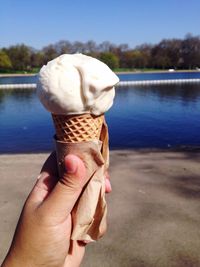 Cropped image of hand holding ice cream cone against lake at park