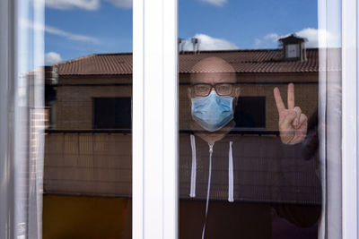 Portrait of man wearing mask showing peace sign seen through window