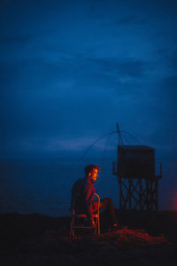 Rear view of man sitting on beach against sky during sunset