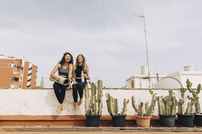 Smiling young female friends sitting together on retaining wall near cactus plants against sky