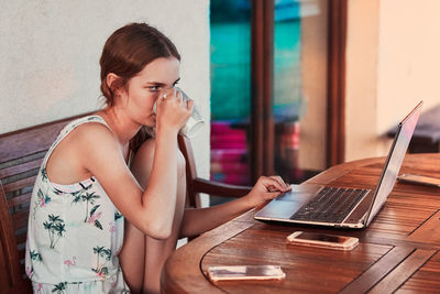 Girl drinking coffee in cup while using laptop on table