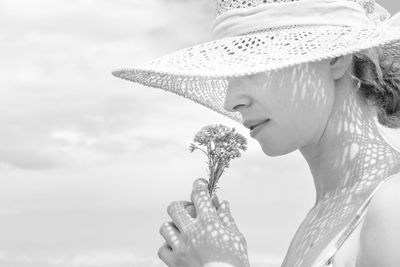 Close-up of woman smelling flower wearing hat against sky