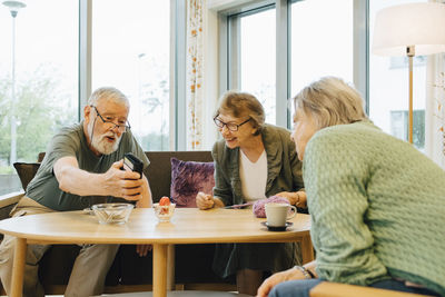 Retired elderly man sharing smart phone with friends sitting at table in nursing home
