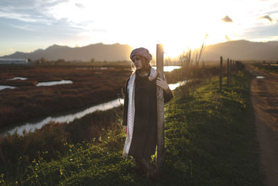 Portrait of young woman standing on field against sky during sunset