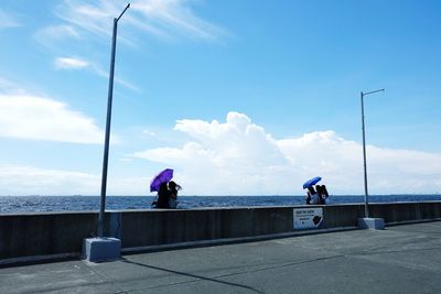 People sitting on retaining wall by sea against blue sky