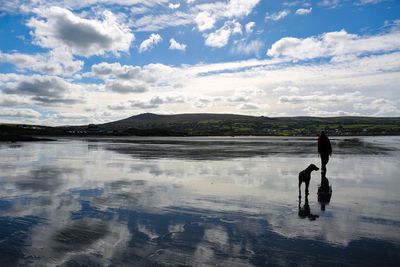 Silhouette man with dog walking on frozen lake against cloudy sky