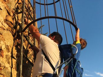 Side view of man with baby boy climbing ladder on wall against sky