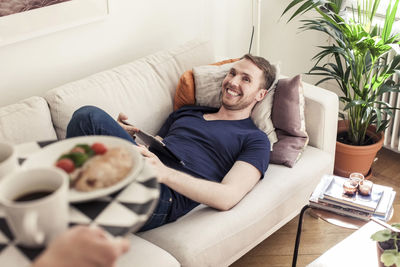 Cropped image of young gay man bringing breakfast for partner lying on sofa