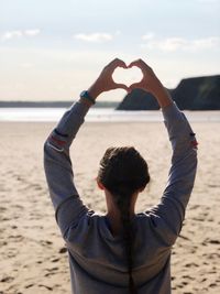 Rear view of woman making heart shape at beach against sky