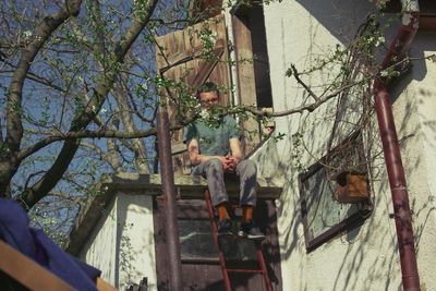 Low angle view of man sitting on ladder by house