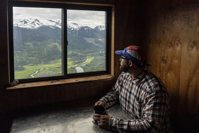 Bearded man in checkered shirt and cap sitting at table with mug of hot beverage and looking at mountains while resting in cozy cabin in british columbia, canada