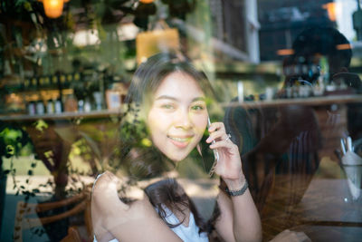 Portrait of woman using mobile phone while sitting at table seen through glass window