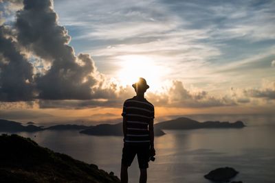 Rear view of man standing at riverbank against cloudy sky during sunset