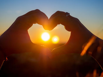 Hand holding heart shape against sky during sunset