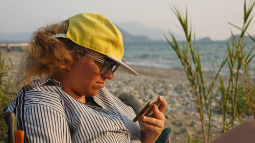 A woman wearing glasses and a baseball cap is sitting on a beach watching a film on her smartphone