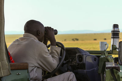 Rear view of man looking through binoculars in vehicle on field