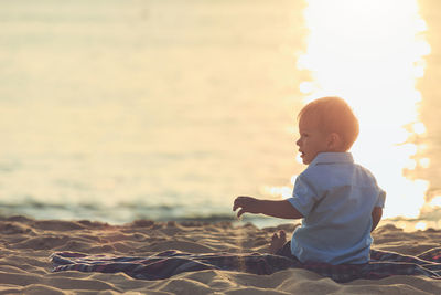 Boy sitting on shore at beach against sky during sunset