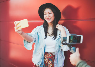 Smiling young woman taking selfie with smart phone while standing against red wall