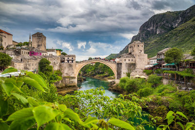Arch bridge over river amidst buildings against cloudy sky