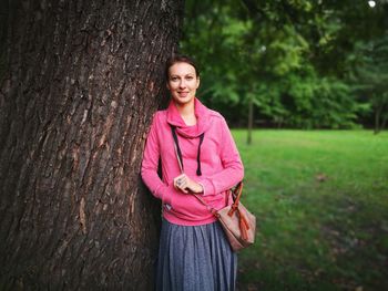 Portrait of smiling young woman standing by tree trunk at park