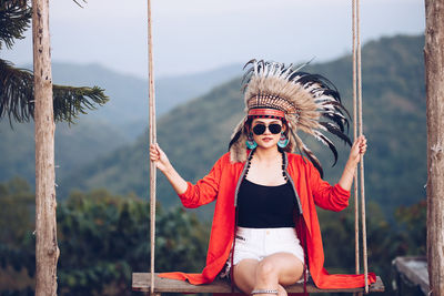 Portrait of young woman wearing headdress while sitting on swing against mountains