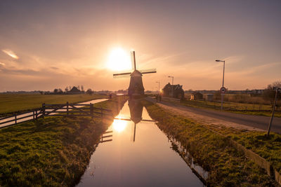 Traditional windmill by canal against sky during sunset