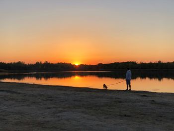 Young man standing with dog at lakeshore during sunset